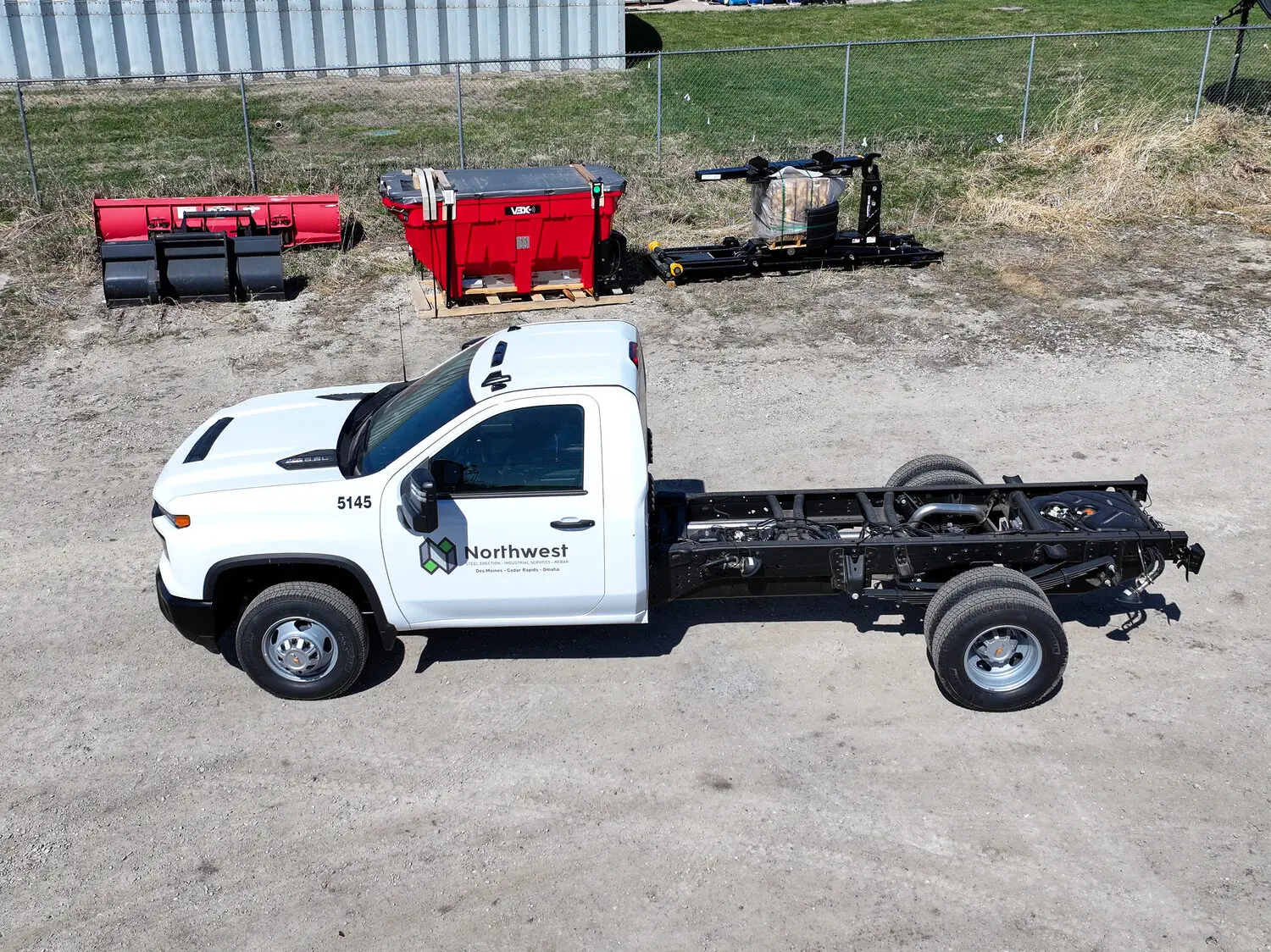 A completed chassis-cab truck photographed from above on the lot, with snow plows and a spreader staged behind it