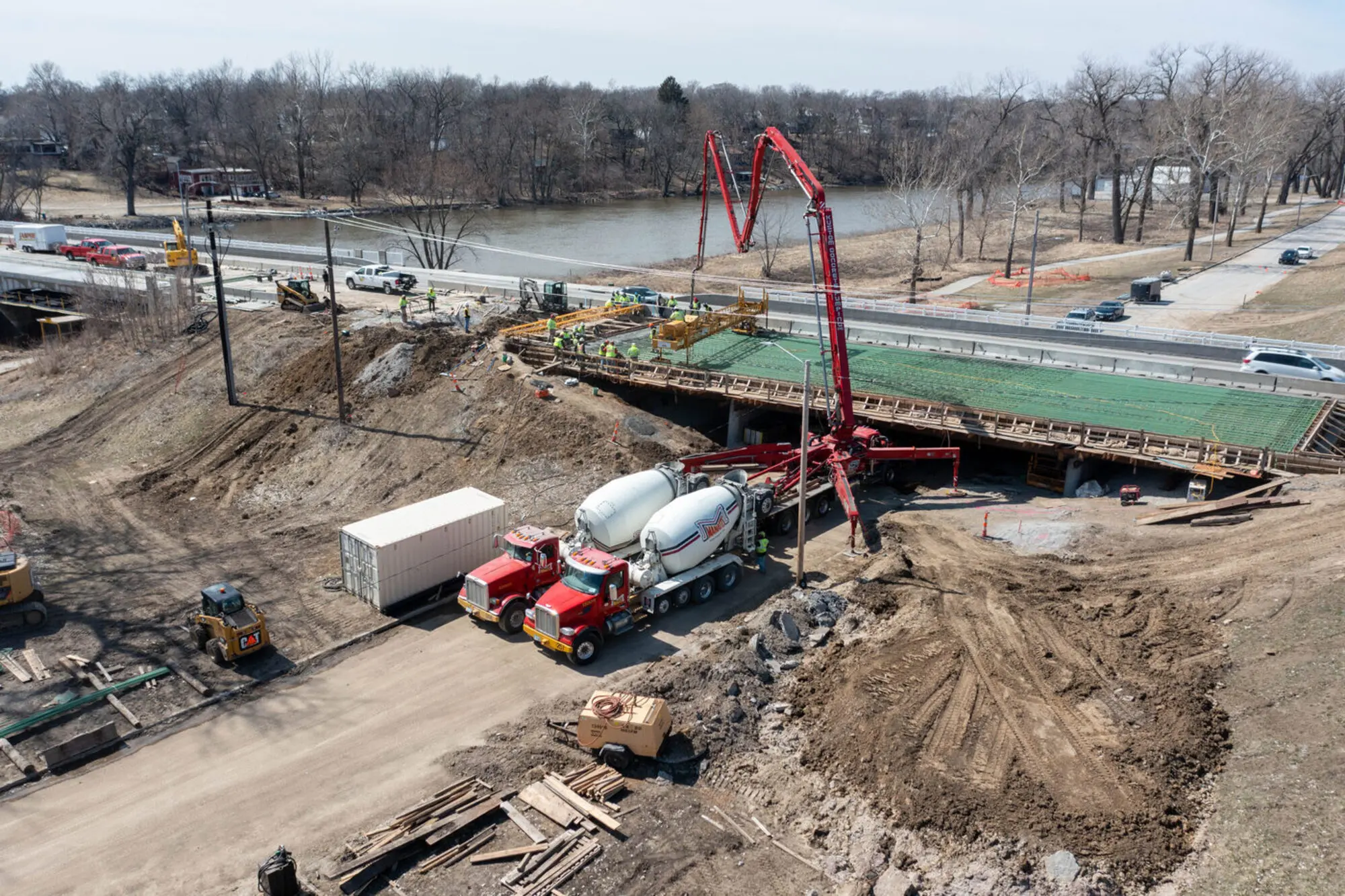 Aerial view of a bridge deck concrete pour: red pump boom extended over the deck, Manatt's mixer trucks queued below, crew working on the rebar mat, river behind