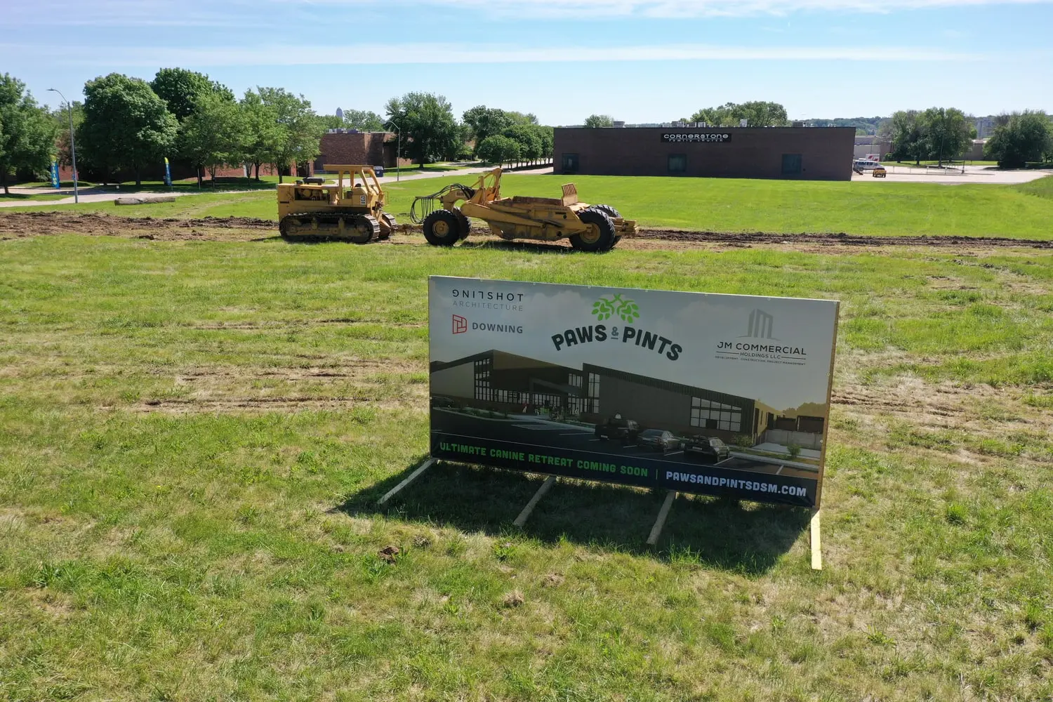 29 May 2021: a Paws & Pints project sign reading Ultimate Canine Retreat Coming Soon, standing in an open grass field with a scraper and dozer working the first cut behind it