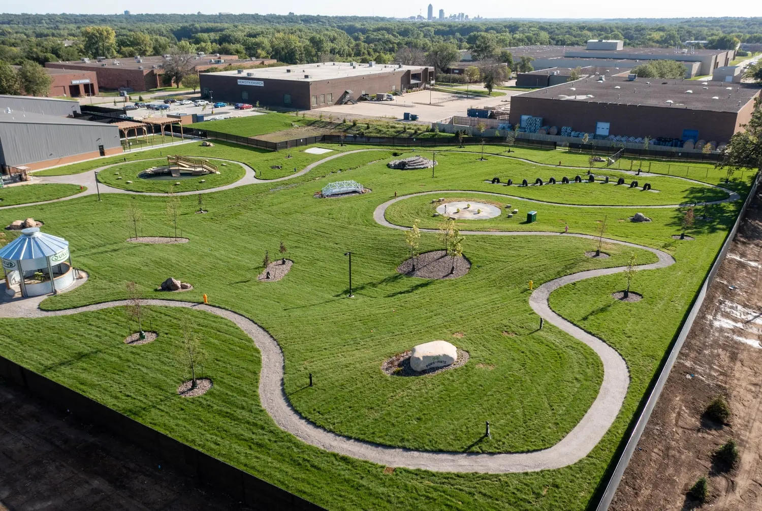 The finished Paws & Pints outdoor dog park in late afternoon light, winding paths and mounded lawn in the foreground, with the Des Moines skyline visible on the horizon