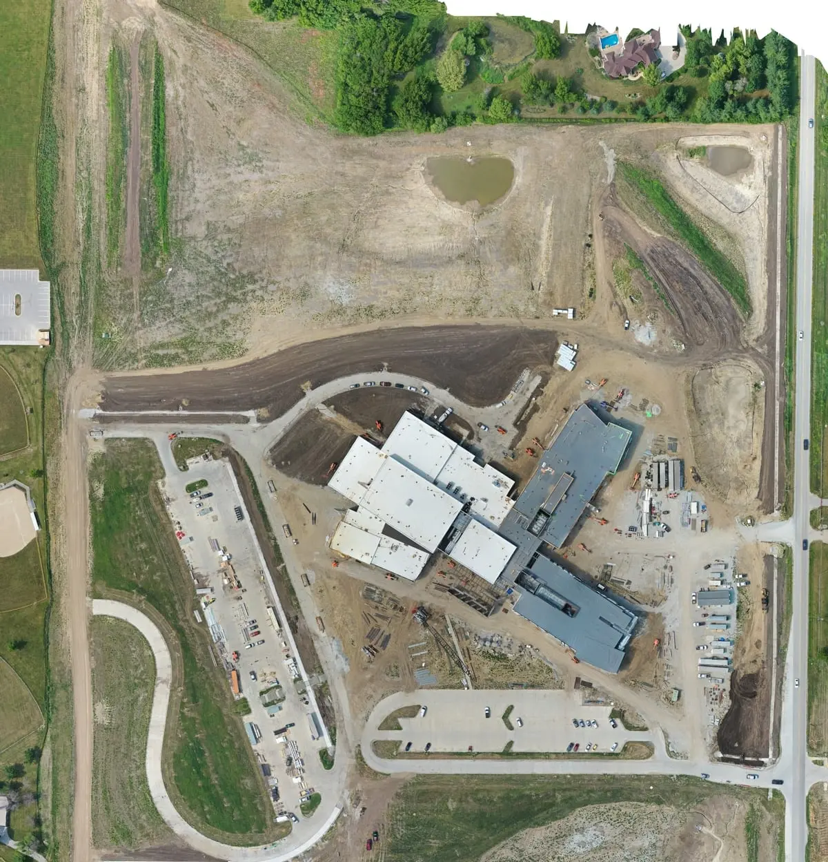 Southeast Polk Middle School site on 27 June 2023: the building shell under roof deck, surrounded by bare fill, unpaved drives and a construction laydown yard